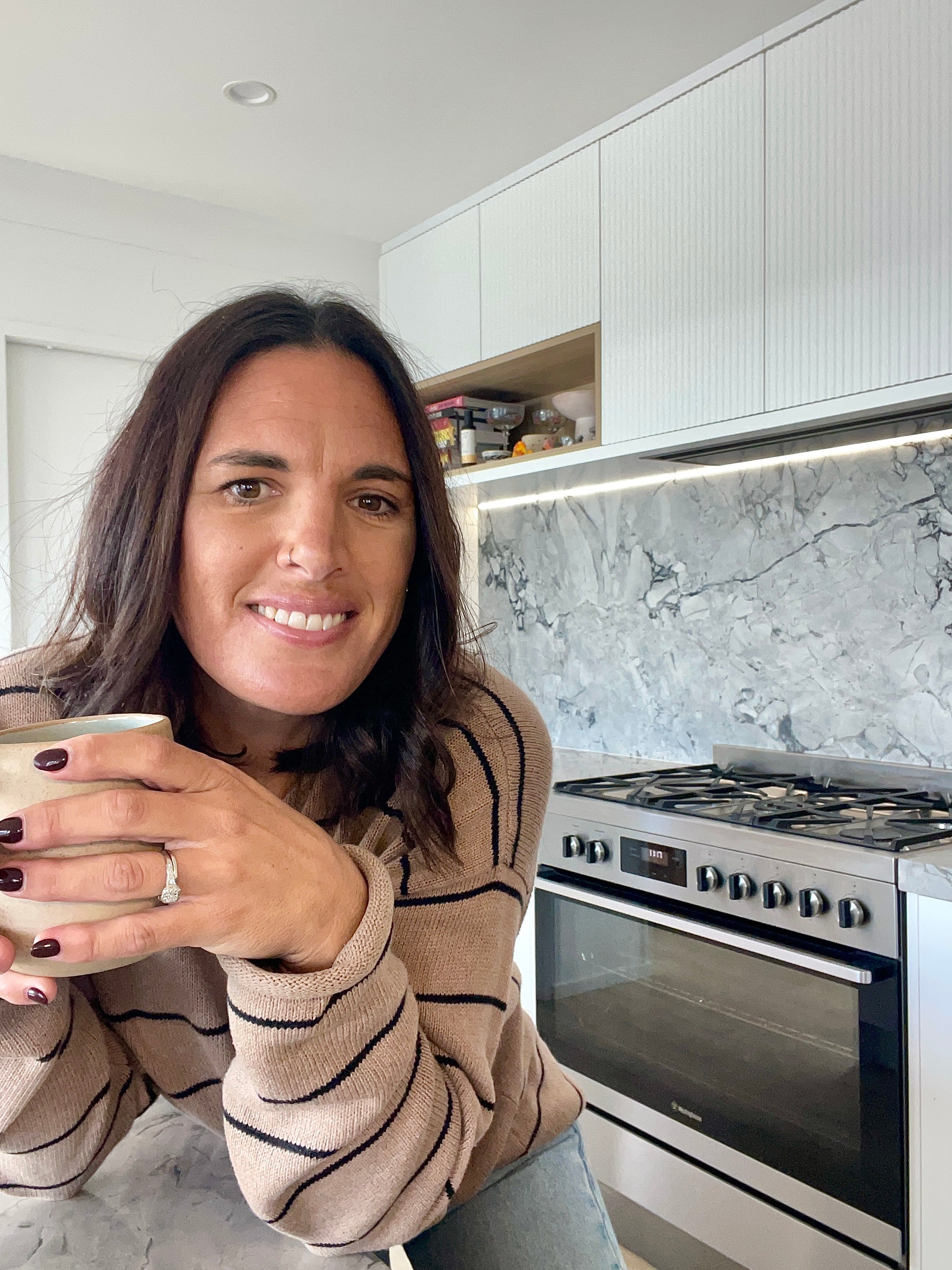 Woman holding a mug in a modern kitchen with white cabinets and marble countertop.